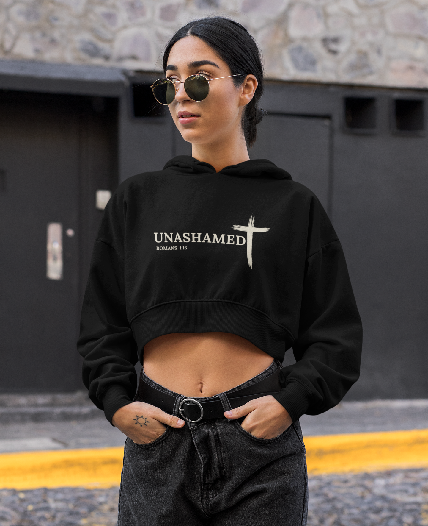 Person wearing a black hoodie with 'UNASHAMED' text and sunglasses, standing against a stone wall.