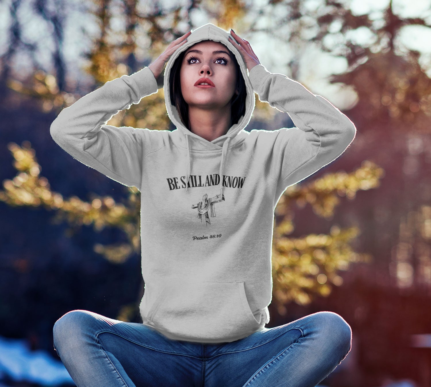 Person wearing a hoodie with text and graphics, sitting outdoors with blurred autumn trees in the background
