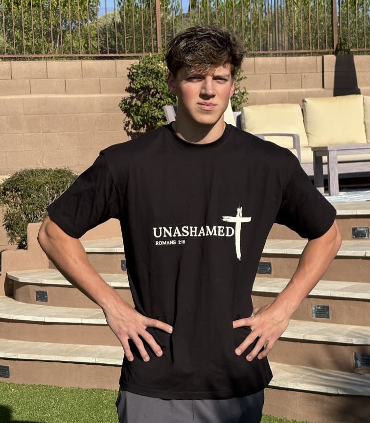 Person wearing a black t-shirt with 'UNASHAMED' text and cross graphic outdoors.