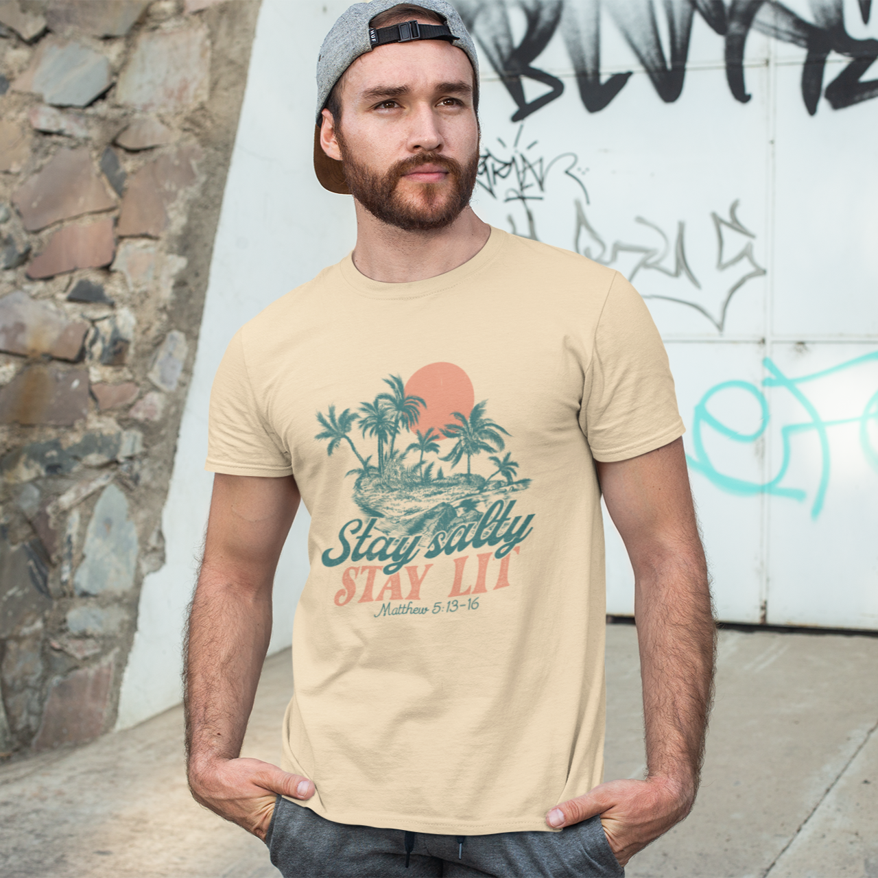 Man wearing a beige t-shirt with a graphic design standing in front of a stone wall and graffiti.